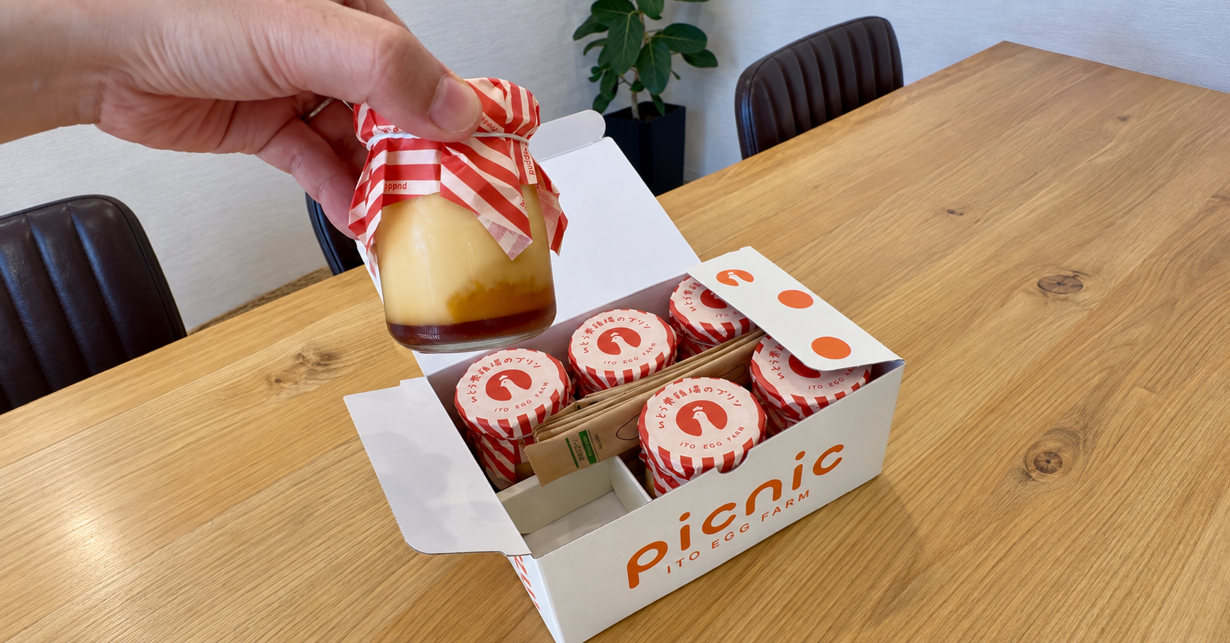 木のテーブルの上で、手に持ったプリンと、同じプリンが並んだ「picnic ITO EGG FARM」の箱を写した写真
