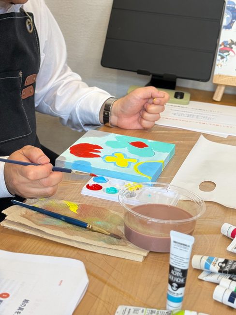 酒井の手元と、水色の下地に赤や黄色の花を描いたキャンバス