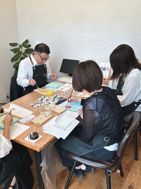 テーブルを囲んで、それぞれのキャンバスに絵を描く酒井とスタッフ