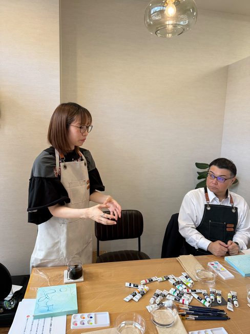 エプロン姿で研修の進め方を説明する講師のChanさんと、席で話を聞く酒井