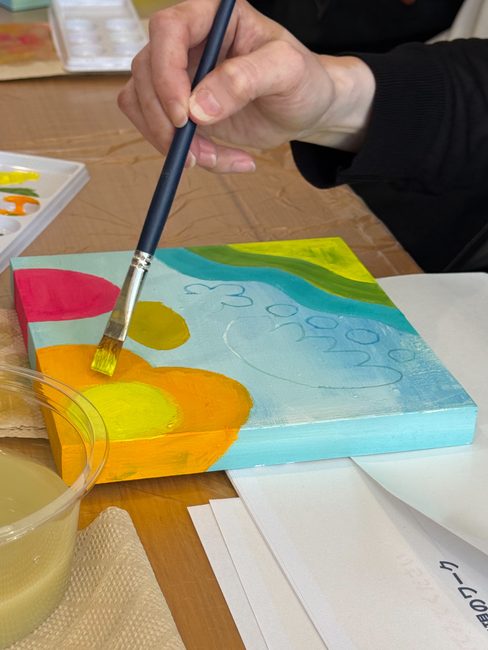 筆を持つ手元と、水色の下地に黄色やピンクの形を描き進めたキャンバス